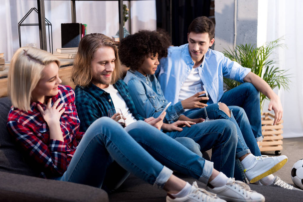 multicultural friends sitting on sofa and looking at smartphone - Foto, Bild