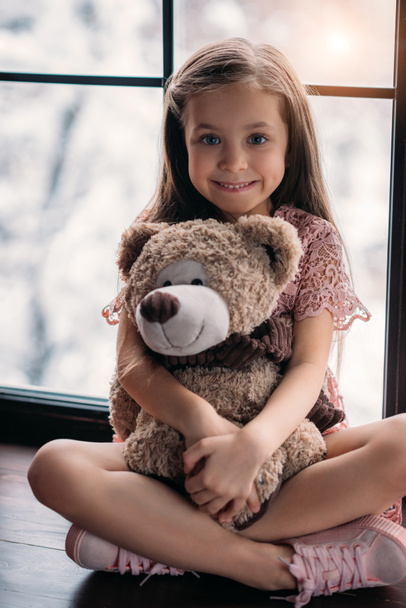 fröhliches kleines Kind auf Fensterbank mit Teddybär - Foto, Bild