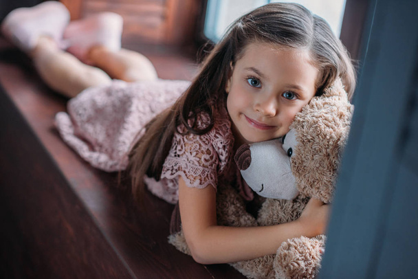 little child embracing teddy bear while lying on windowsill - Фото, изображение