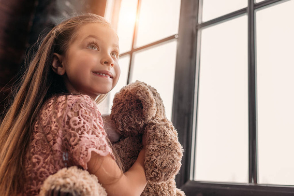 happy little child playing with teddy bear on windowsill - Foto, afbeelding