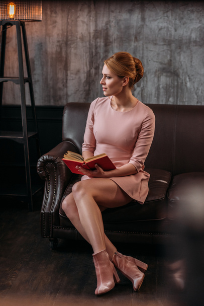 mujer joven reflexiva con libro sentado en el sofá y mirando hacia otro lado
 - Foto, imagen