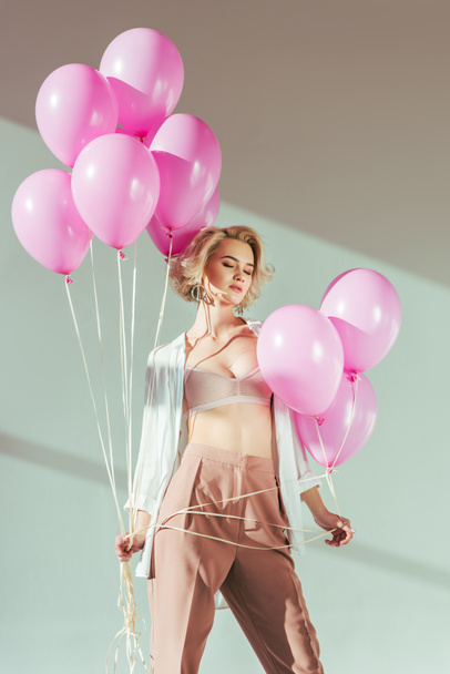 соблазнительная молодая женщина в модной одежде и лифчике позирует с розовыми шариками на сером
  - Фото, изображение