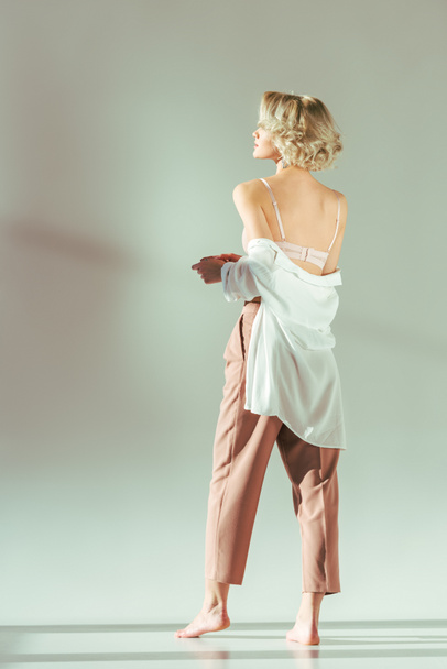 back view of barefoot blonde girl in pink bra, shirt and pants standing and looking away on grey - Photo, Image