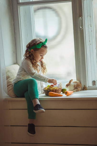Pretty happy child girl is sitting on window sill with her friend little colorful rabbit, Easter holiday concept - Fotografie, Obrázek