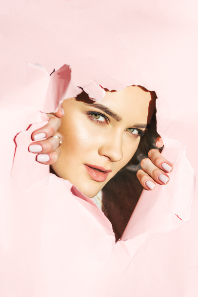beautiful young woman looking at camera through torn pink paper  - Фото, зображення