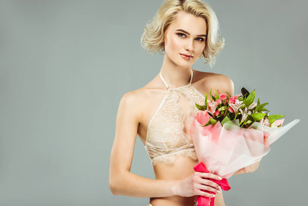 attraktive blonde Mädchen in Spitze-BH mit Blumenstrauß, isoliert auf grau - Foto, Bild