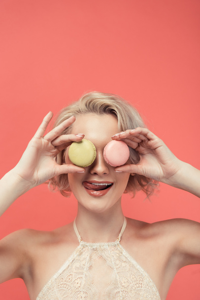 beautiful girl licking lips and holding two macarons in front of eyes, isolated on red - Photo, Image