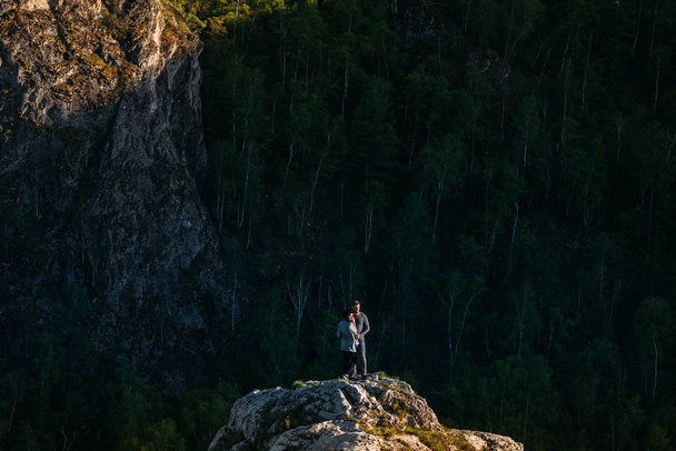 Loving couple at sunset standing on a cliff. Guy and girl hugging at sunset. Couple travels. Lovers in nature. Man and woman watching the sunset. Lovers at sunset. Travel in the mountains. Relations - 写真・画像