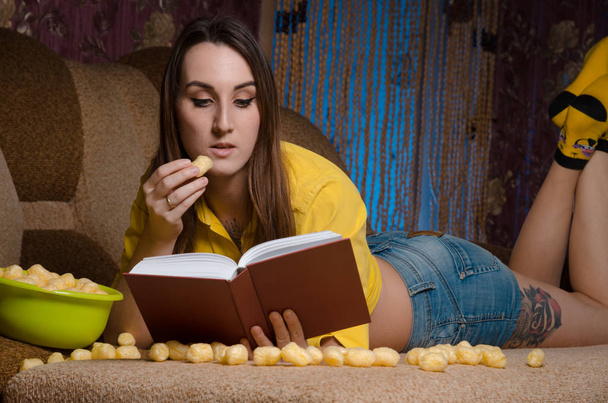 the girl on the sofa reading a book and eating Cheetos - Foto, Imagen