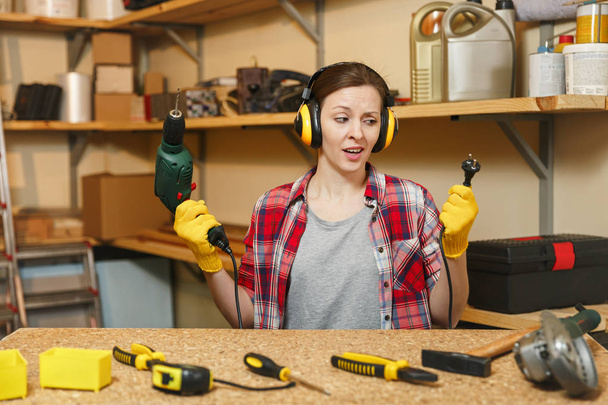 Perplexed young woman in plaid shirt gray T-shirt noise insulated headphones yellow gloves working in carpentry workshop at wooden table place with piece of wood, different tools, power drill, wire. - Photo, Image