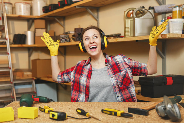 Молодая женщина в клетчатой рубашке, серая футболка, шумоизоляционные наушники, желтые перчатки, расправляющие руки, работающая в столярной мастерской на деревянном столе с куском дерева, различные инструменты. Копирование пространства
 - Фото, изображение
