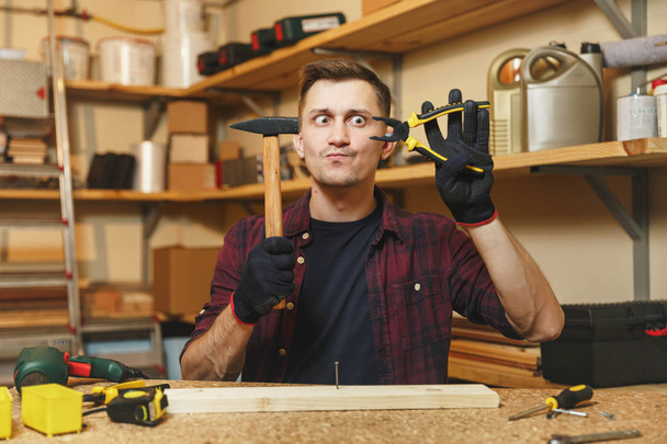 Crazy fun mad caucasian young man in plaid shirt, black T-shirt, gloves hammering nails with hammer, working in carpentry workshop at wooden table place with piece of wood, pliers, different tools. - 写真・画像