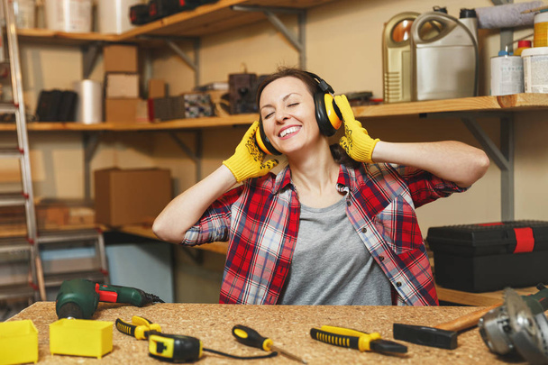European young woman in plaid shirt, gray T-shirt, noise insulated headphones, yellow gloves listening music, working in carpentry workshop at wooden table place with piece of wood, different tools. - Photo, Image