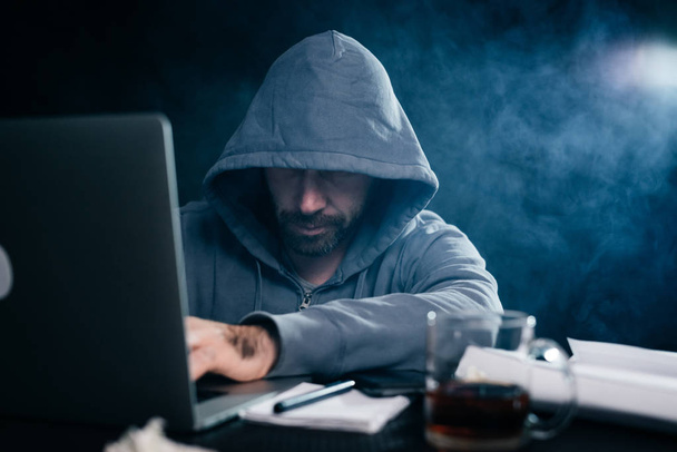 A computer hacker is typing on a laptop in a smoky room. Dark background - Fotoğraf, Görsel