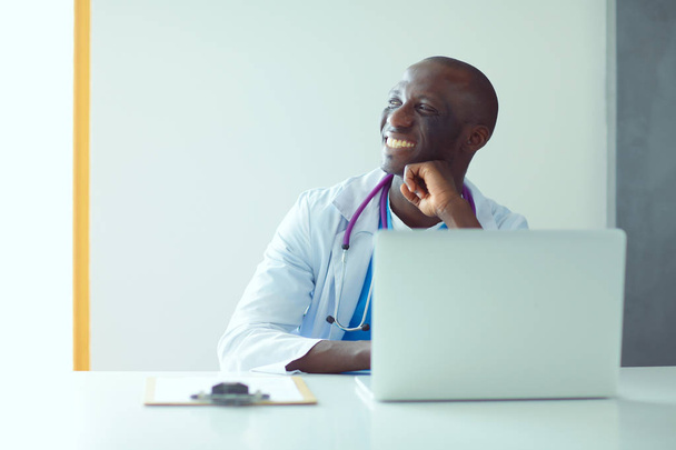 Portret van een jonge mannelijke arts die een headset draagt terwijl hij een computer gebruikt aan een bureau in de kliniek. Dokter.. - Foto, afbeelding