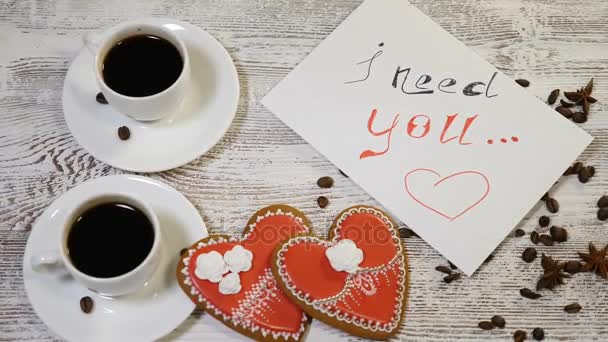 Vista dall'alto di un messaggio I need You e due tazze di caffè con biscotti allo zenzero a forma di cuore su un tavolo di legno. Concetto di relazione amorosa. San Valentino. Girato in 4K
 - Filmati, video