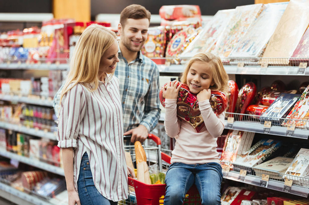 Smiling little girl sitting on a shopping cart and choosing candy with her parents at the supermarket - Foto, Imagem