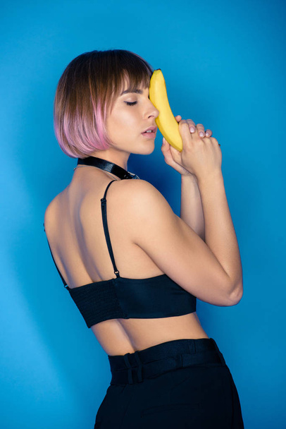 stylish girl in crop top touching face with banana isolated on blue - Valokuva, kuva