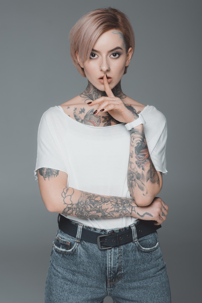 hermosa chica tatuada haciendo un gesto de silencio y mirando a la cámara aislada en gris
 - Foto, Imagen
