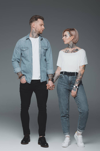 full length view of beautiful stylish young tattooed couple holding hands and looking at each other on grey - Photo, Image