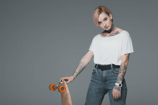 beautiful young tattooed woman standing with skateboard and looking at camera isolated on grey - Fotó, kép