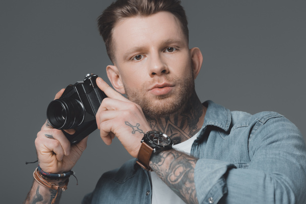 handsome young man with tattoos holding camera and looking at camera isolated on grey - Fotoğraf, Görsel