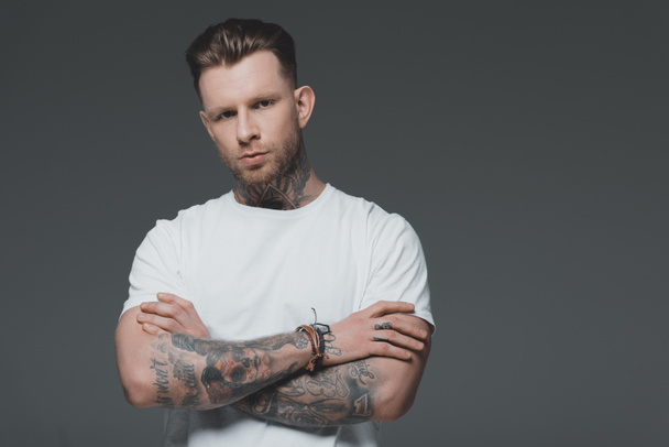 portrait of handsome young tattooed man in white t-shirt standing with crossed arms and looking at camera isolated on grey - Foto, Imagem
