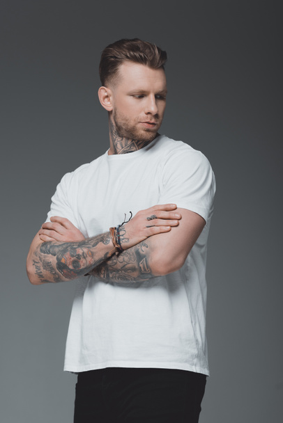 handsome stylish young tattooed man in white t-shirt standing with crossed arms and looking away isolated on grey - Фото, зображення