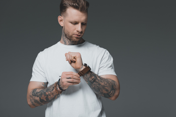 bonito elegante jovem tatuado homem no branco t-shirt vestindo relógio de pulso isolado no cinza
 - Foto, Imagem