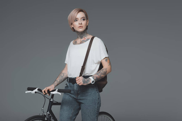 beautiful tattooed girl with backpack standing with bicycle and looking away isolated on grey  - Fotó, kép