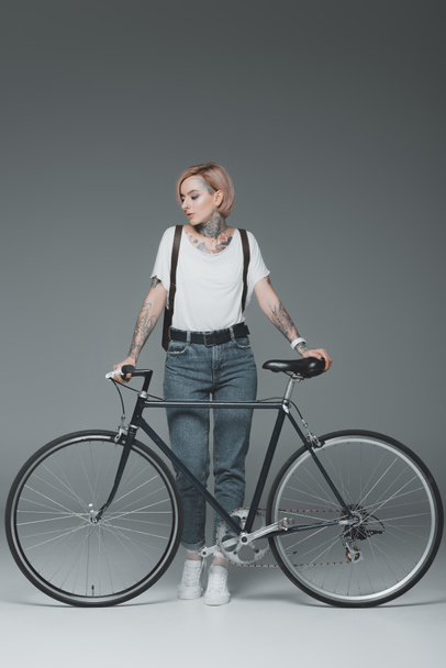beautiful stylish girl with tattoos standing with bicycle isolated on grey - Fotó, kép