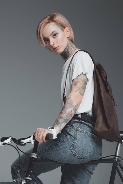 beautiful tattooed girl sitting on bicycle and looking at camera isolated on grey  - Foto, Bild