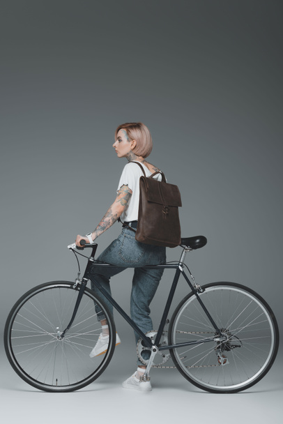 stylish young tattooed woman with backpack sitting on bike and looking away on grey - Photo, Image