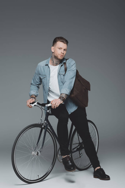 стильный молодой человек с татуировкой сидит на велосипеде и смотрит в камеру на сером
 - Фото, изображение