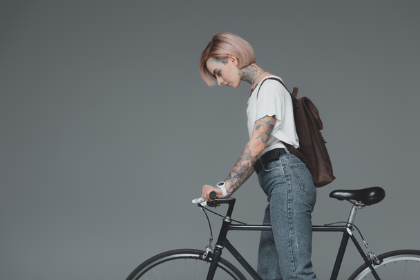 vista lateral de mujer joven con mochila de pie con bicicleta aislada en gris
 - Foto, Imagen