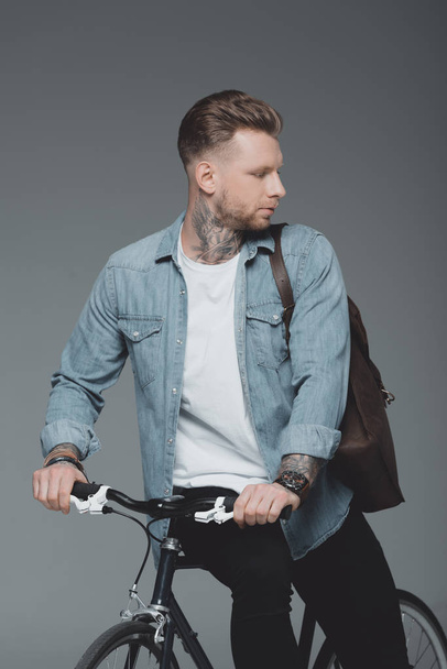 stylish young man with shoulder bag sitting on bicycle and looking away isolated on grey - Foto, immagini