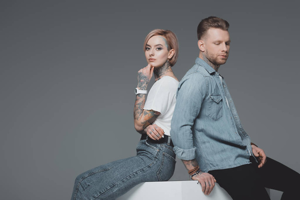 stylish young tattooed couple sitting back to back and looking at camera isolated on grey  - Photo, Image