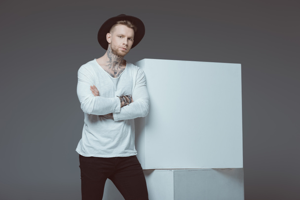 stylish young tattooed man in black hat standing with crossed arms and looking at camera isolated on grey - Foto, Imagem