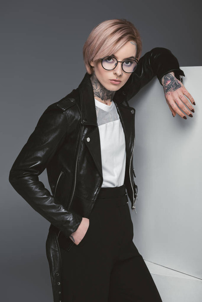 stylish young tattooed woman in leather jacket leaning at white cubes and looking at camera isolated on grey - Fotografie, Obrázek