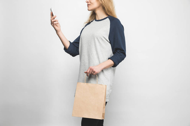 Schnappschuss einer Frau in stylischem Langarm auf Weiß mit Einkaufstasche und Smartphone - Foto, Bild