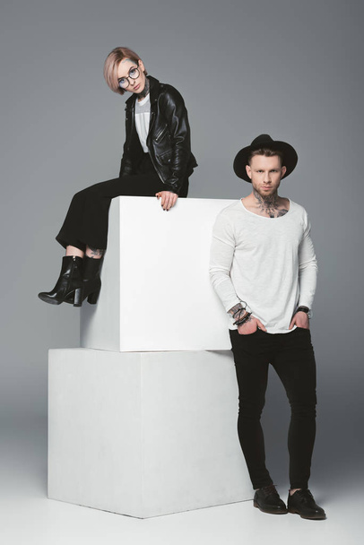 full length view of stylish young tattooed couple posing together and looking at camera on grey  - Valokuva, kuva