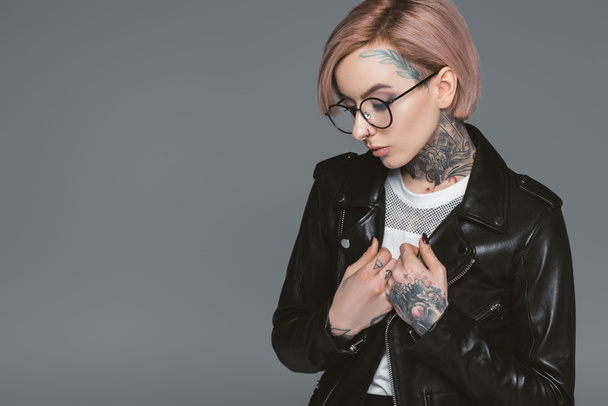 fashionable tattooed girl in eyeglasses and black leather jacket, isolated on grey - Foto, imagen