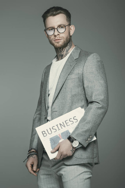 stylish businessman in eyeglasses and suit holding business newspaper, isolated on grey - Fotó, kép