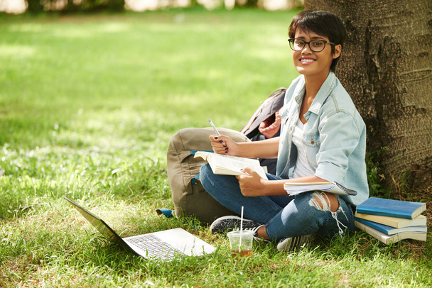 Estudante asiático atraente sentado no gramado verde e tomando notas necessárias, retrato de comprimento total
 - Foto, Imagem