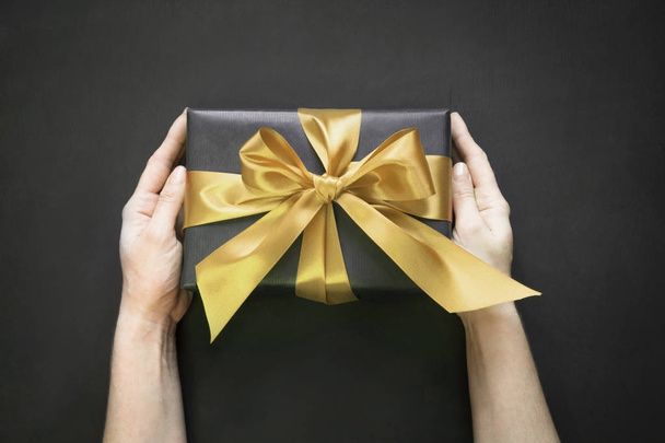 Gift box wrapped in black paper with gold ribbon in female hand on black surface. Top view. - 写真・画像