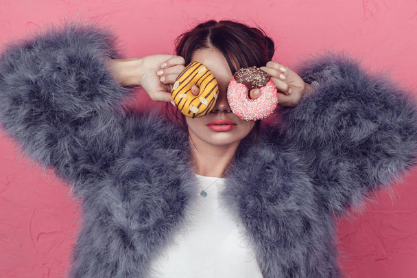 Beautiful girl in a fur coat with donuts in hands posing on a pink background. - Foto, Imagem