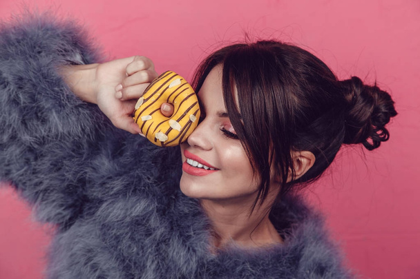 Beautiful girl in a fur coat with donuts in hands posing on a pink background. - Фото, зображення