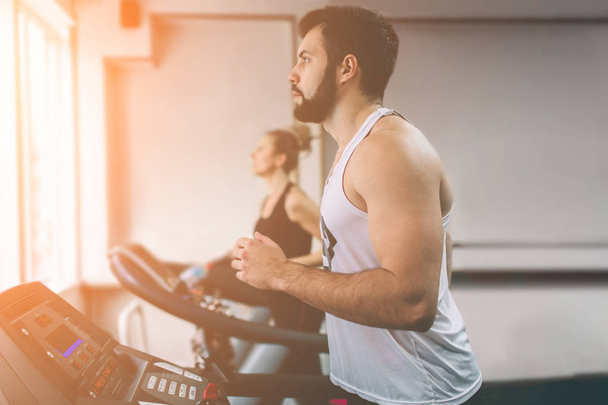 Young man in sportswear running on treadmill at the gym. Muscular bearded athlete during workout . Close up of young athletic female model trains at the indoor fitness center. - 写真・画像