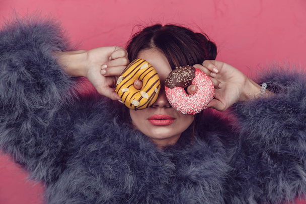 Beautiful girl in a fur coat with donuts in hands posing on a pink background. - Foto, Imagen