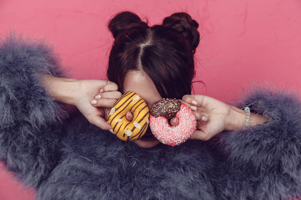 Beautiful girl in a fur coat with donuts in hands posing on a pink background. - Valokuva, kuva